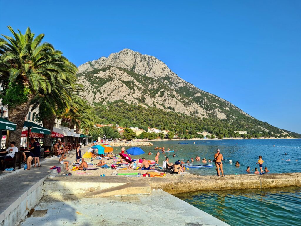 Beachgoers enjoying a sunny day on the pebble beach with palm trees and mountain backdrop in Gradac, Croatia
