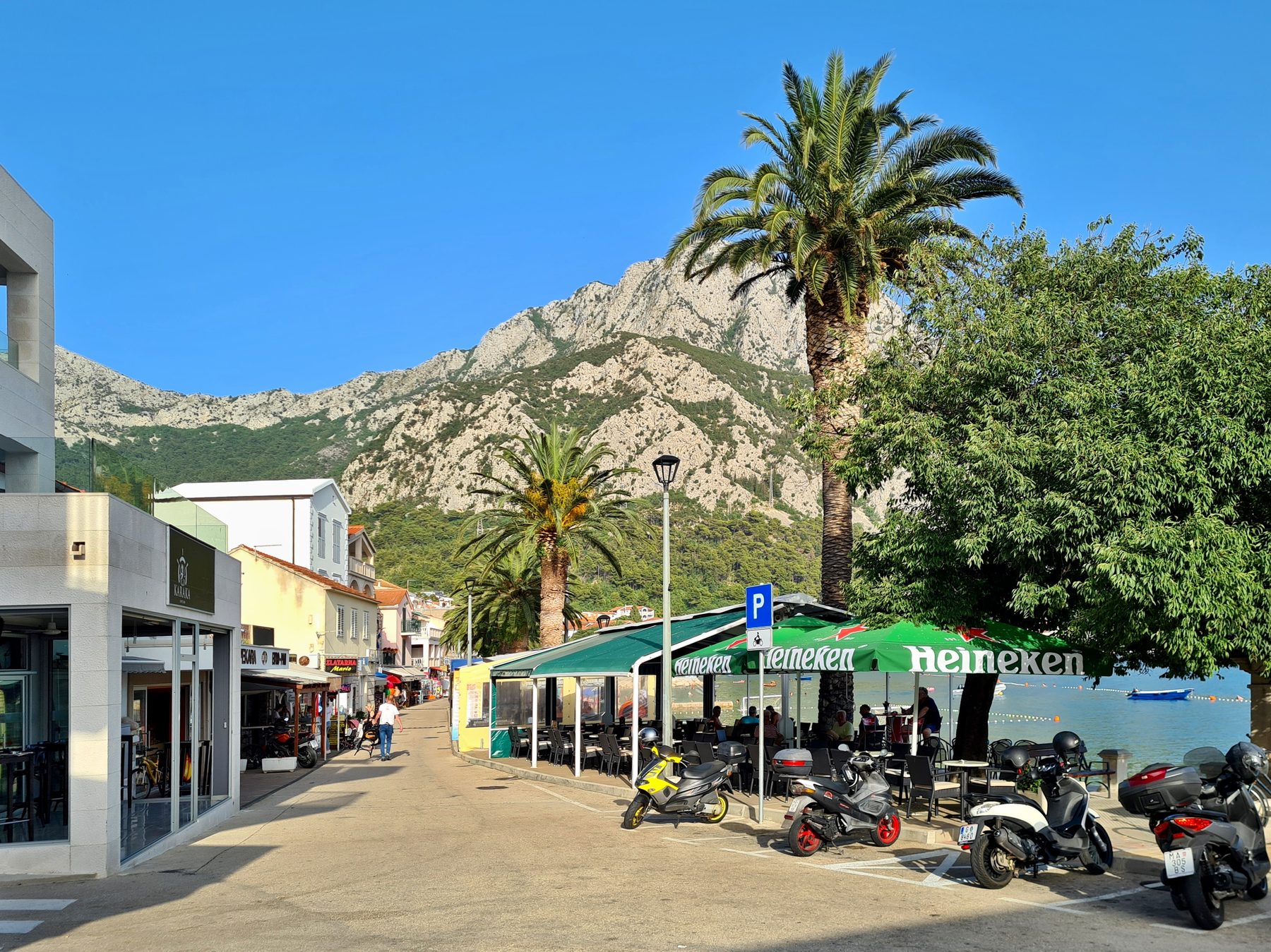 Street with outdoor cafe by the sea, palm trees, scooters, and mountains in Gradac, Croatia