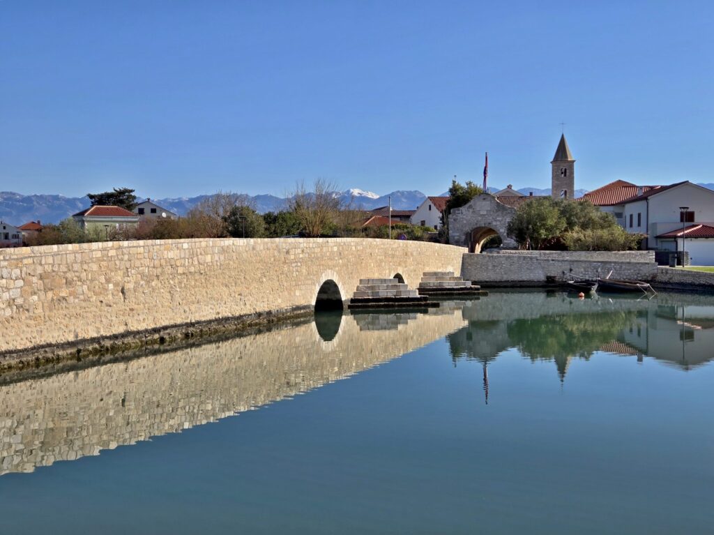 Stone bridge over calm water with reflections and historic buildings in Nin, Croatia under clear sky