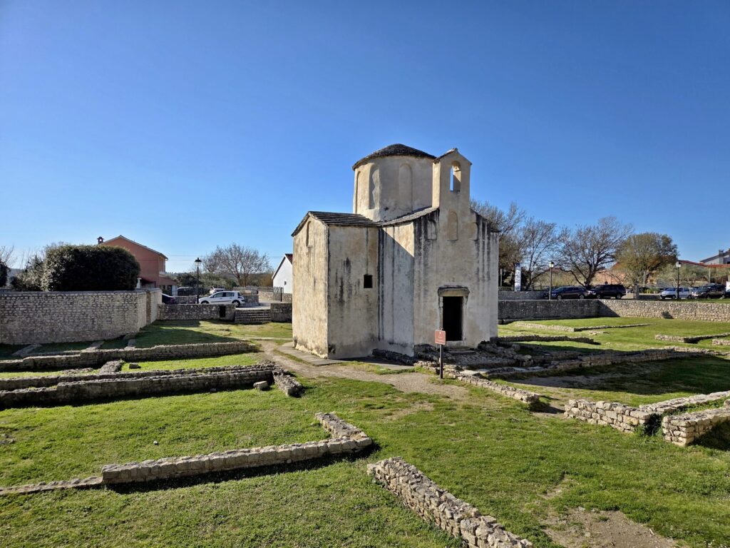 Ancient stone church surrounded by grass and low stone walls ruins in Nin, Croatia under clear blue sky