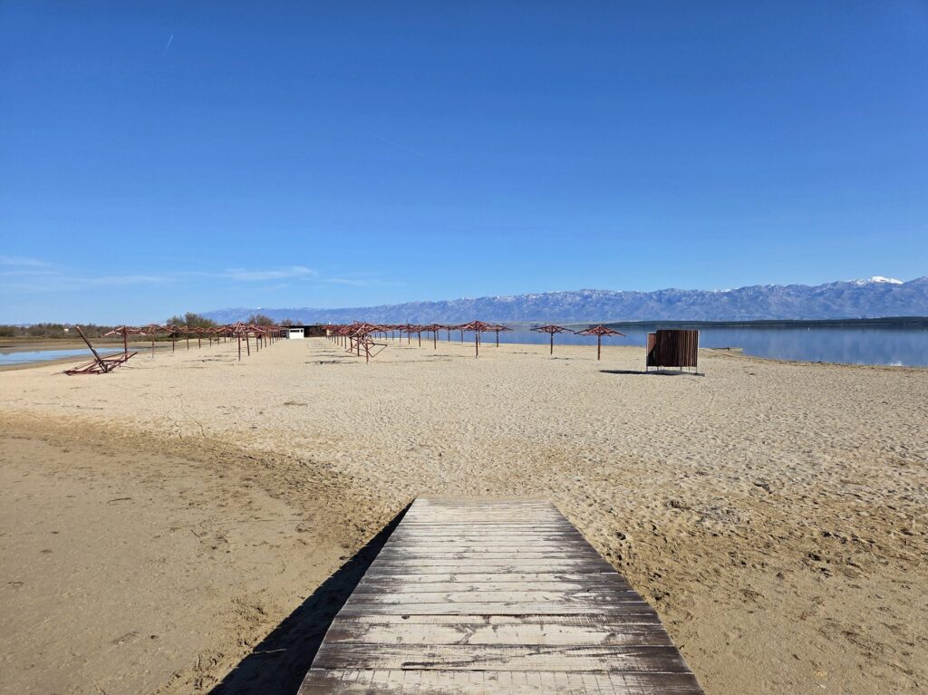Sandy beach with wooden walkway and empty sunshades under blue sky near Nin, Croatia