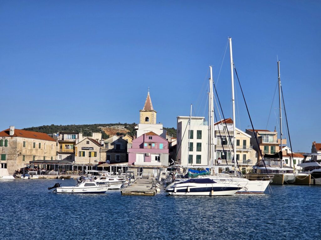 Boats docked in harbor with historic stone buildings and church tower in Pirovac, Croatia