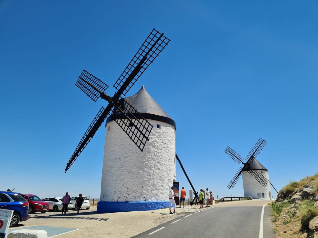 Large white windmills beside the road in Consuegra, Spain with visitors nearby.
